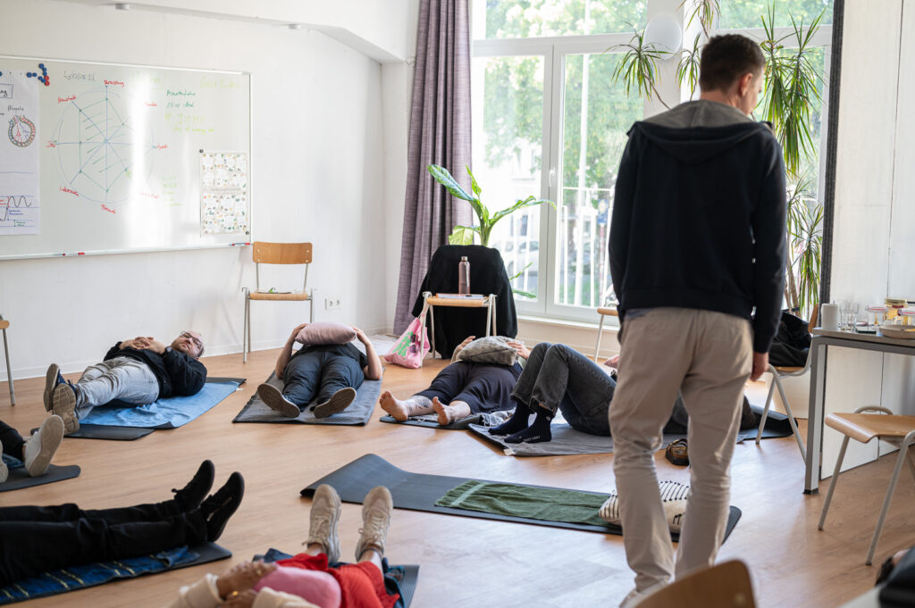 Breathwork für Anfänger Teilnehmende liegen auf Yogamatten mit Kissen auf ihren Bäuchen, während Paul eine Atemübung anleitet.