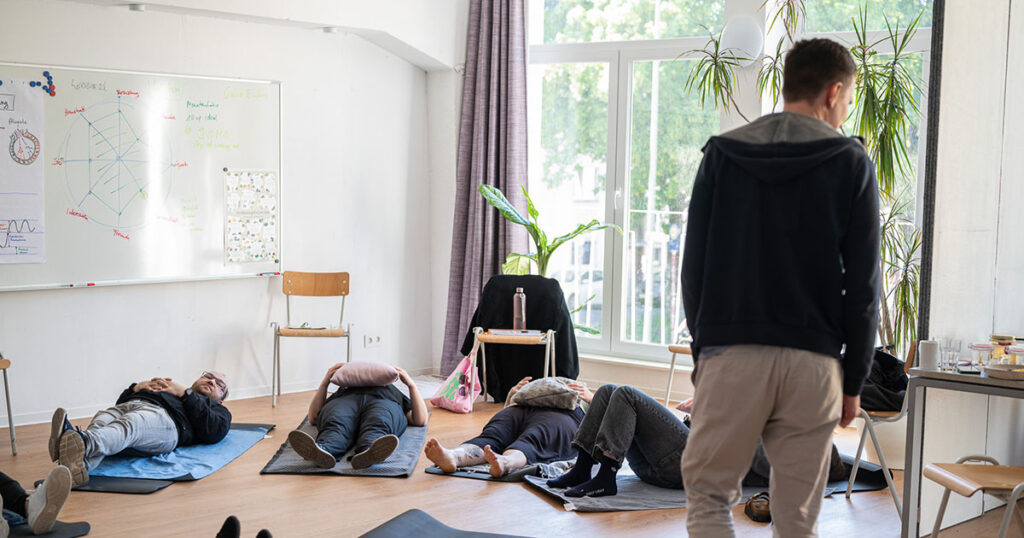 Teilnehmende liegen auf Yogamatten mit Kissen auf ihren Bäuchen, während Paul eine Atemübung anleitet.